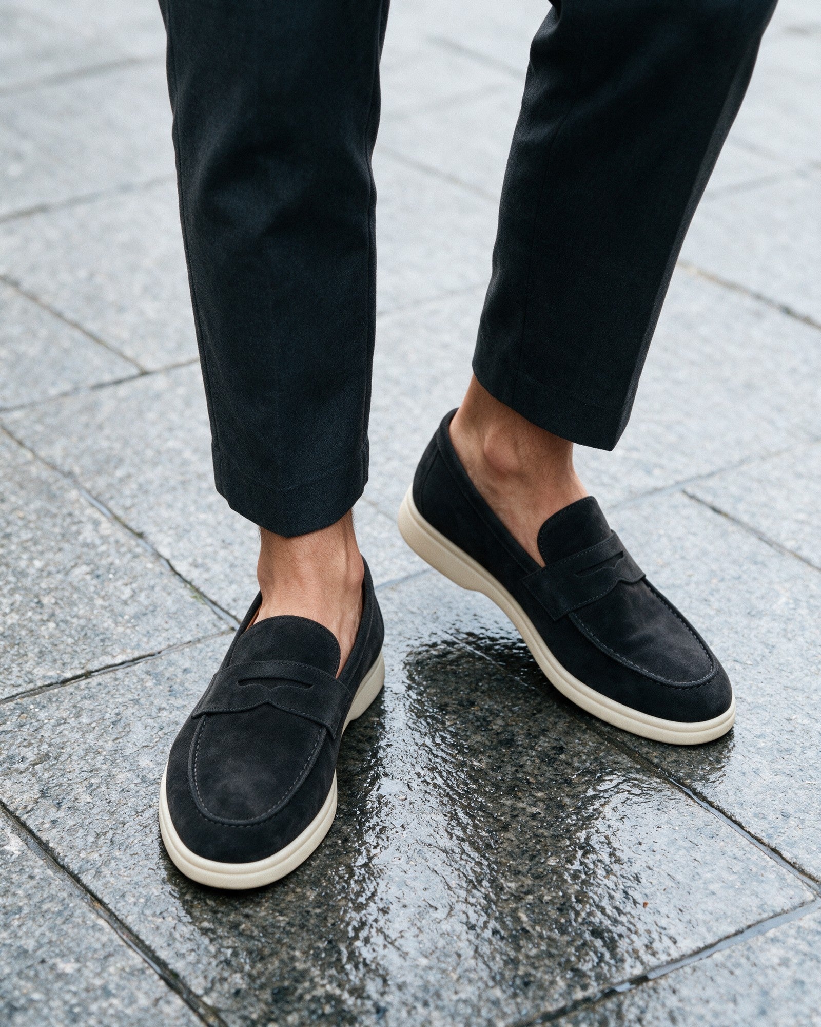 Lorvano Suede Penny Loafers in black suede with cream sole worn on wet granite paving