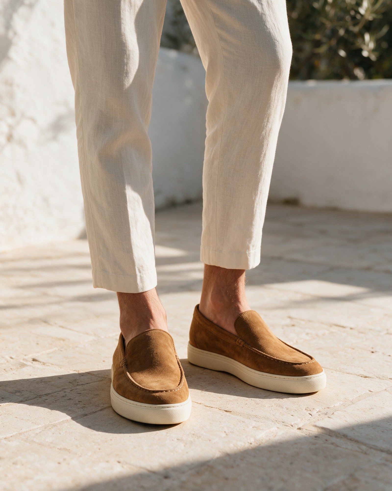 Lorvano Suede Loafers in camel tan suede worn with cream linen trousers on a limestone terrace